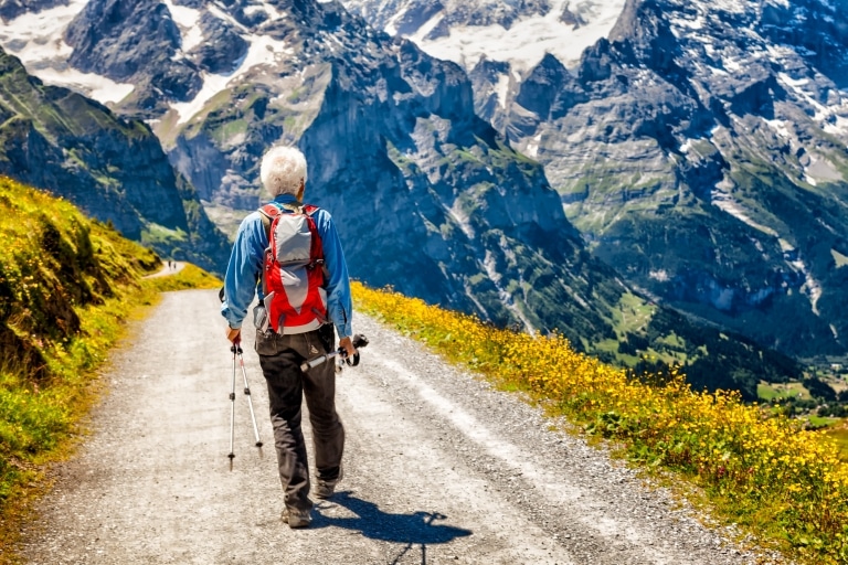 Mann mit Wanderbekleidung und Wanderstöcken geht einen Weg entlang, im Hintergrund zeigt sich eine Bergkulisse