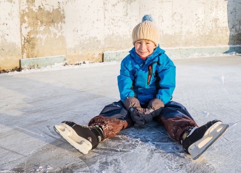 Symbolbild Eislaufen in Landeck: kleiner Junge mit Schlittschuhen sitzt auf der Eislaufbahn