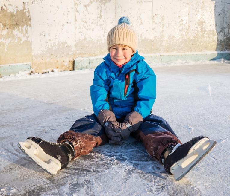 Symbolbild Eislaufen in Landeck: kleiner Junge mit Schlittschuhen sitzt auf der Eislaufbahn